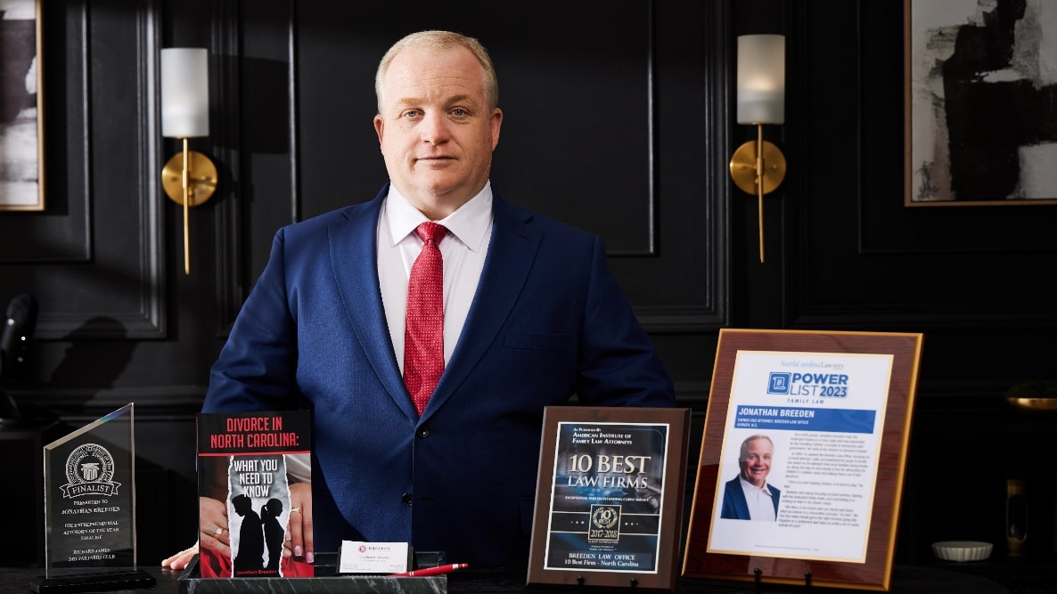 Attorney Jonathan Breeden standing next to his awards to the 10 Best Law Firms from the American Institute of Family Law Attorneys, the North Carolina Law Power List 2023, and Entrepreneurial Attorney of the Year Finalist - as well as his book "Divorce in North Carolina: What You Need to Know"