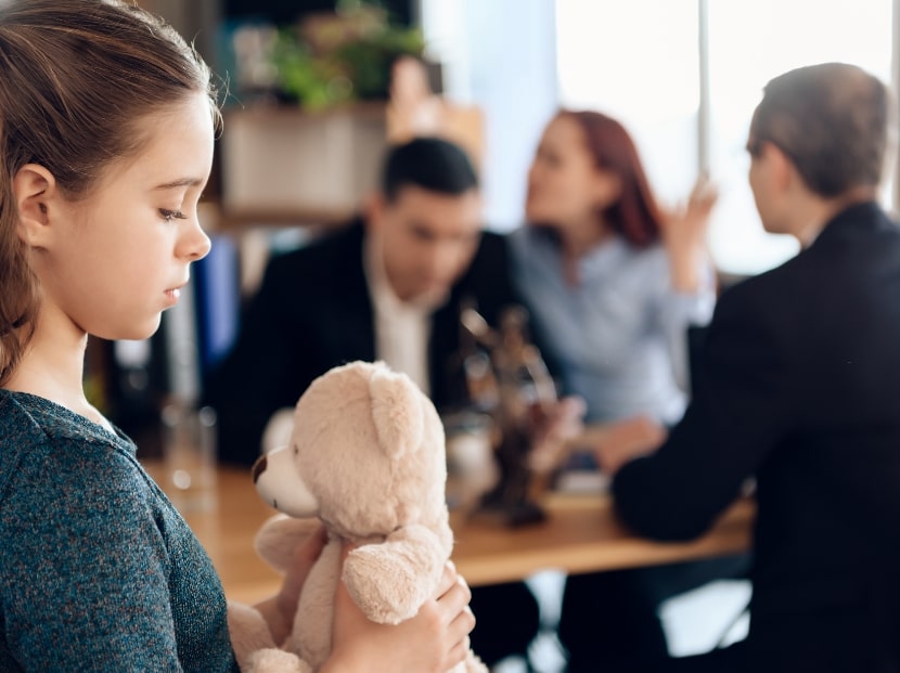 Sad young girl holding stuffed animal as parents argue in the background